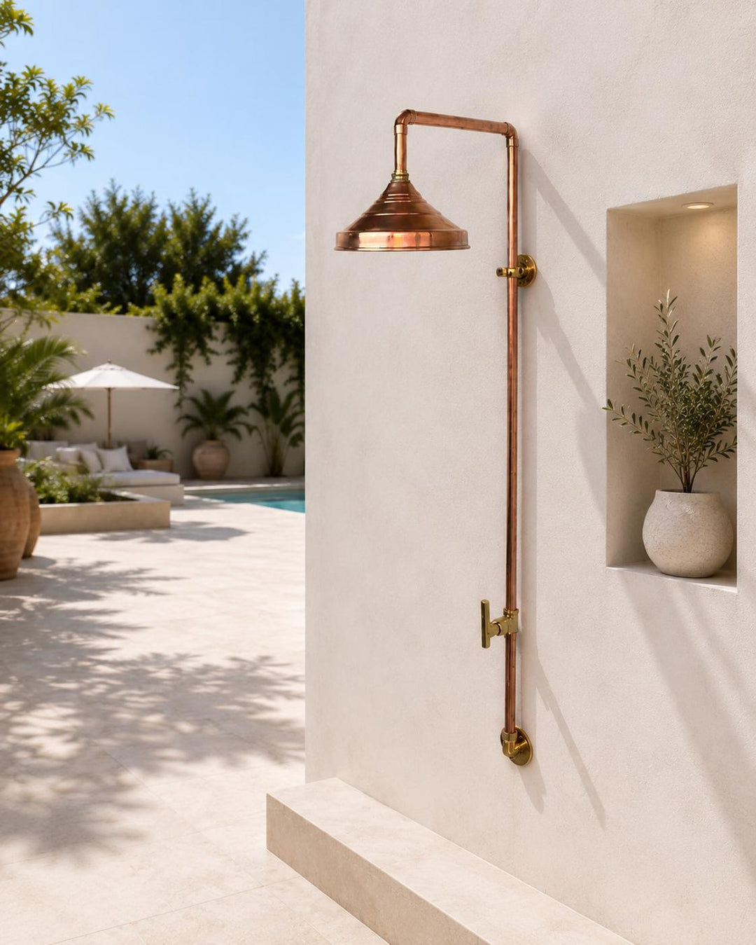 A copper and brass wall-mounted rain shower head with a long vertical pipe, installed on a beige stucco wall in a modern outdoor patio setting.