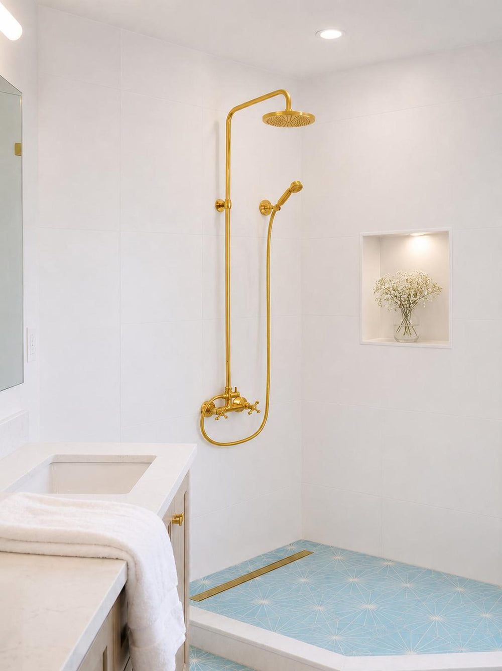 A modern bathroom featuring a prominent gold shower fixture with a rain head and handheld shower head mounted on a white tiled wall. The shower base has light blue patterned floor tiles, and a white vanity with a marble countertop is visible in the foreground.