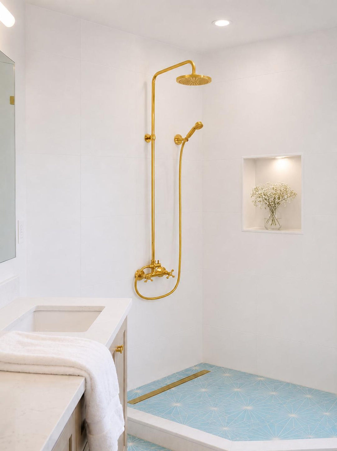 A modern bathroom featuring a prominent gold shower fixture with a rain head and handheld shower head mounted on a white tiled wall. The shower base has light blue patterned floor tiles, and a white vanity with a marble countertop is visible in the foreground.