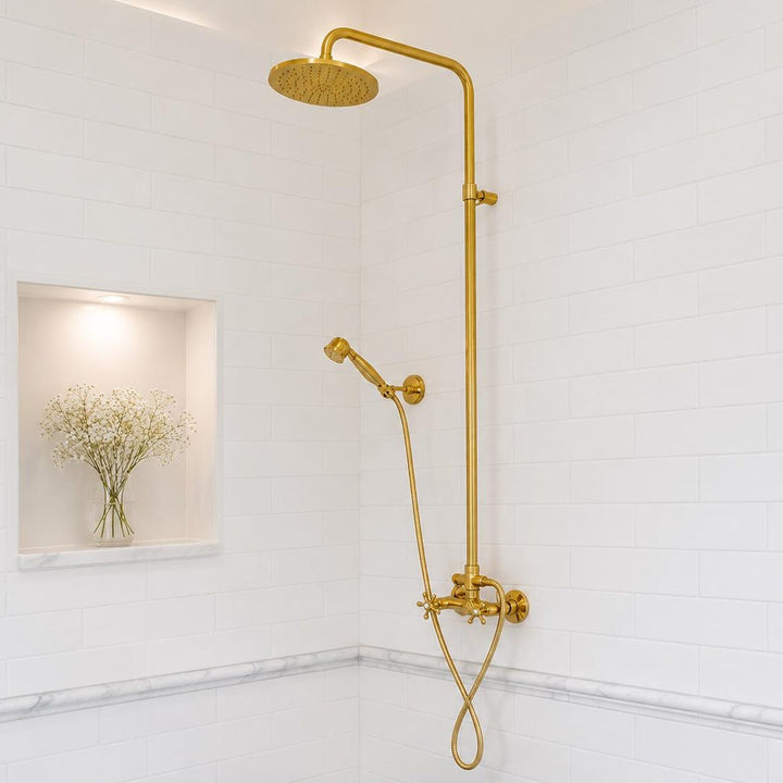 A modern bathroom corner featuring a gold-finished shower system with a round rain showerhead, adjustable vertical bar, handheld shower, and brass valves. The bathroom has white subway tiles, a recessed niche with baby&
